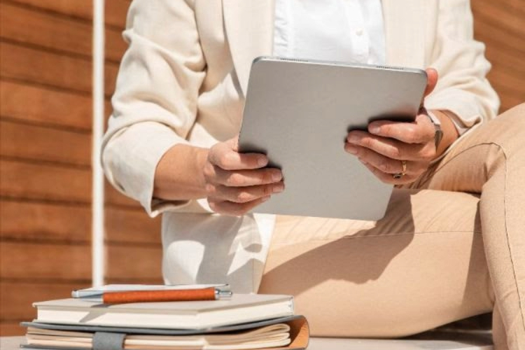 This image depicts a person in business casual clothing holding a tablet and sitting next to a stack of notebooks. This picture is meant to represent a frontline recruiter working through recruitment strategy for 2024.